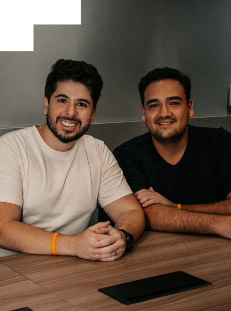 Dois homens sorrindo sentados lado a lado em uma mesa de reunião, usando pulseiras laranjas. O ambiente é moderno e iluminado, com clima descontraído.